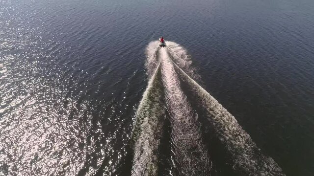 Aerial view of people ride on jetski on the city pond. People on a jet ski sail off into the distance. There are houses and trees on the shore. On the other bank, forest and hills. Summer sunny day.
