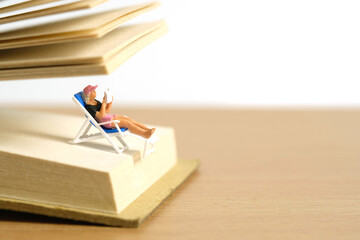 Miniature people toy figure photography. Girl relaxing seat at beach chair while reading book