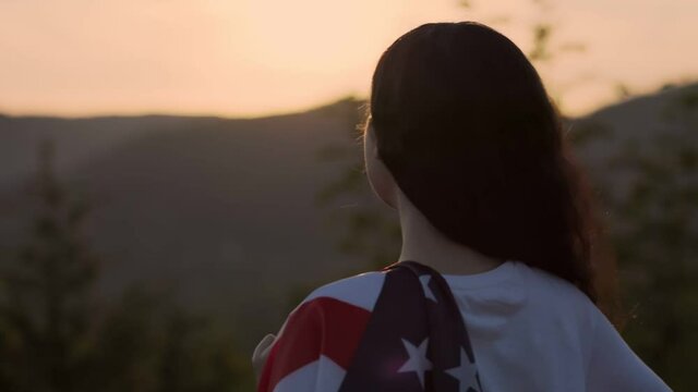 Independence Day. A young woman with an American flag draped over her shoulder, against the background of the sunset sky. Slow motion. Back view. Dolly shot. The concept of patriotism.