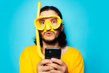 Portrait of young surprised man in yellow, wearing diving mask with snorkel, using smartphone on...