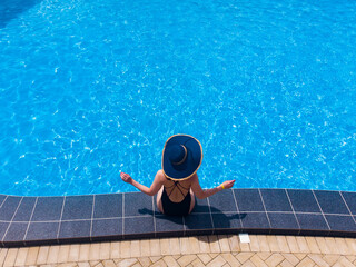 Fototapeta na wymiar woman sitting on the edge of the pool wearing straw hat, from above view, aerial top view from drone, summer relaxation in hotel