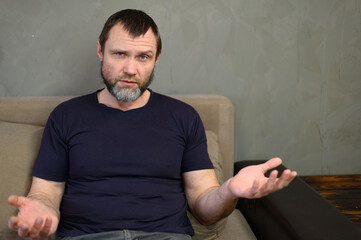Portrait of adult caucasian serious man with beard talking to camera while sitting on sofa at home