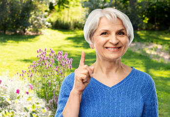 gardening and old people concept - portrait of smiling senior woman in blue sweater pointing finger up over summer garden background