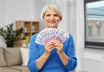 savings, finances and people concept - smiling senior woman holding fan of five hundred euro money banknotes over home living room background
