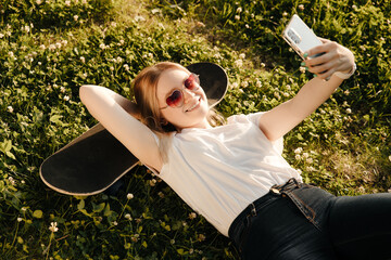 the girl lies on the grass with her head on a skateboard and takes a selfie