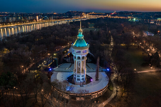 Budapest, Hungary - Aerial view of illuminated Margaret Island Open-Air Stage (Margitszigeti Szabadteri Szinpad) water tower at dusk with blue and golden sky and downtown of Budapest at background