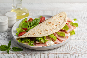 Piadina romagnola, traditional italian flatbread with soft cheese, ham and lettuce. On a white wood table.
