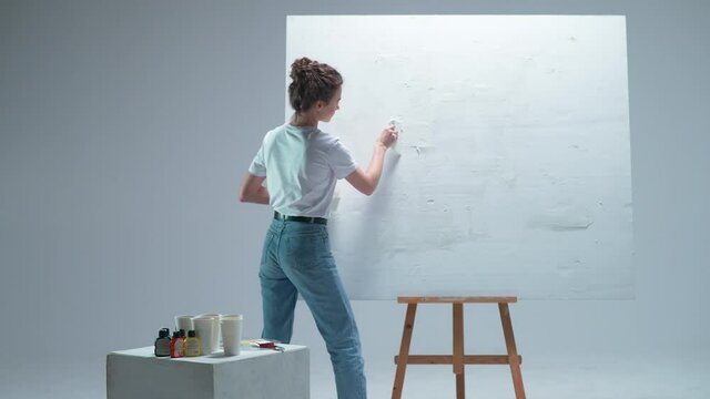 Back view, female artist applies a layer of white primer on a large canvas in a white room, a talented artist preparing to draw, 4k slow motion.