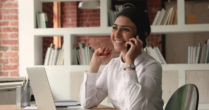 Smiling young indian woman giving professional consultation to client by phone call, sitting at table with computer in modern office. Happy millennial business lady discussing working issues distantly