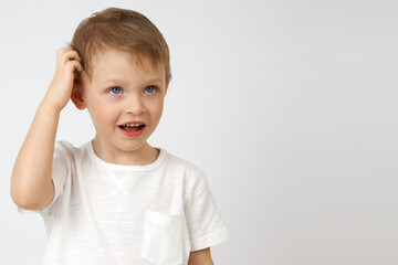 Little boy with blue eyes is deep in thought, looking up and scratching his head against a white background. Solving a difficult task by a child