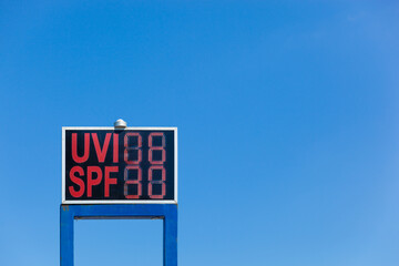 Electronic display with UV radiation warning and info about recommended SPF with red letters, bright blue summer sky in background