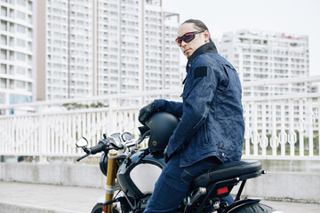 Fototapeta na wymiar Serious man in denim jacket and sunglasses sitting on motorcycle and turning back to camera
