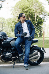 Fototapeta na wymiar Handsome man in jeans and denim jacket leaning on motorcycle and drinking cup of take-out coffee after long ride