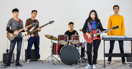 Group shot of five teenage musicians playing the music and singing a song together. Young students play bass guitar, electric guitar, drum, and keyboard. Concept of a teenager playing with friends