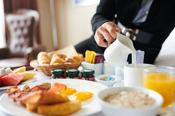 Woman sitting on hotel bed and pouring fresh milk in cup of coffee when eating tasty breakfast