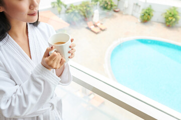 Cropped image young woman in white bathrobe drinking big mug of morning coffee when standing at hotel window and looking outside