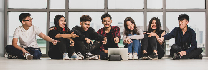 Group of seven young male and female teenagers in casual clothes sitting on the floor and pointing fingers at the tablet. College student calling video online with a friends to making a conversation