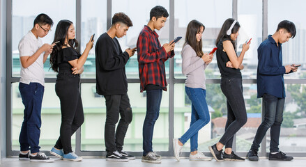 Full-length body portrait shot of a group of seven young male and female teenagers standing and using smartphones and tablets while waiting for a queue. Concept of technology addicted in modern life
