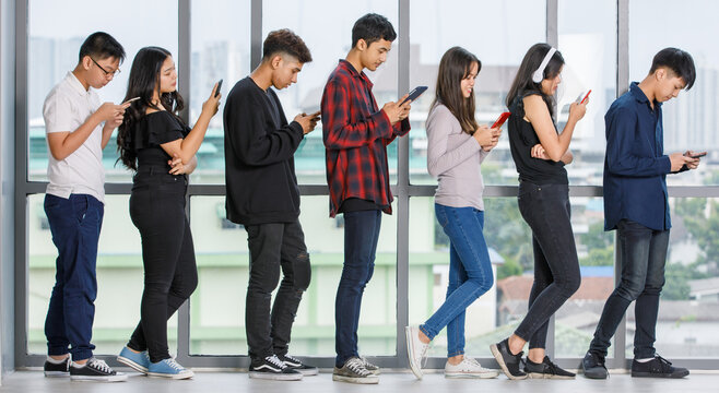Full-length body portrait shot of a group of seven young male and female teenagers standing and using smartphones and tablets while waiting for a queue. Concept of technology addicted in modern life