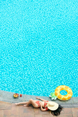 Young woman in bikini relaxing by swimming of spa hotel, she is sunbathing next to inflatable ring, bowl of fruits and glass sweet cocktail