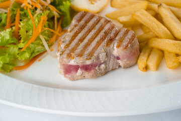 Tuna steak grilled with french fries salad of carrots, parsnips, lettuce and lemon on white plate on white background.