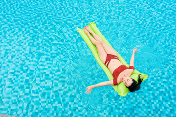 Slim young woman in red bikini relaxing on inflatable mattress floating in swimming pool of spa resort