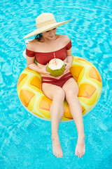 Young woman in straw hat sitting on inflatable ring in swimming pool and drinking coconut cocktail