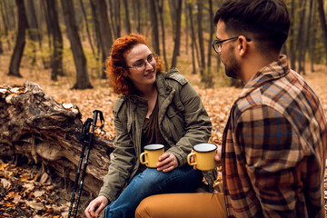 Young couple with backpack sitting on collapsed trunk resting and drinking tea after hiking in forest.