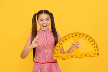 winking teen girl hold school math tool protractor ruler on yellow background show ok, education