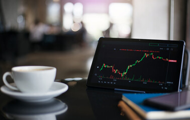 Focus on digital tablet screen showing graph of investment in stock or currency with white coffee cup, notebook and smart phone on table