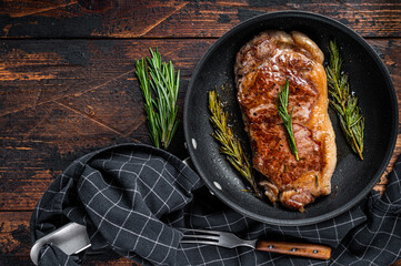 Grilled new york strip beef meat steak in a pan with herbs. Dark wooden background. Top view. Copy space