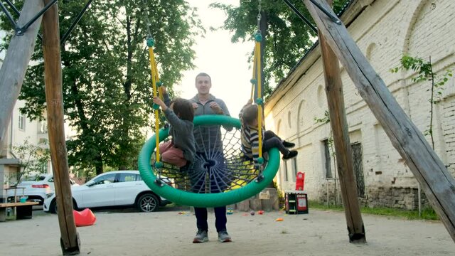 Father shakes his children on swing under tree in sun. close-up. child laughs and rejoices. Parentr and kids ride on rope swing on an oak branch in forest. Family fun in park, in nature outdoors