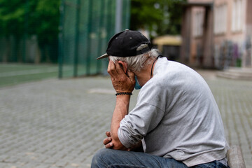 An elderly gray-haired man in a black cap sits sadly on a bench