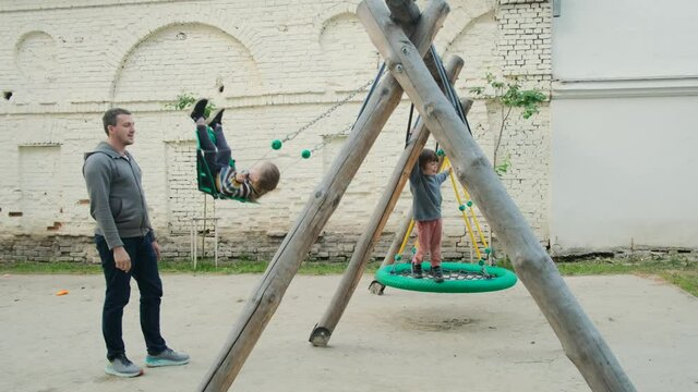 Father shakes his children on swing under tree in sun. close-up. child laughs and rejoices. Parentr and kids ride on rope swing on an oak branch in forest. Family fun in park, in nature outdoors