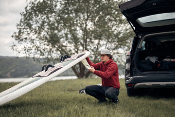Windsurfer and camper packing and unpacking from a car's roof rack in nature.