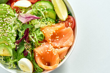 Delicious veggie bowl with salmon, avocado, cucumber, cherry tomatoes and mix salad in a white plate. Isolated on grey background.