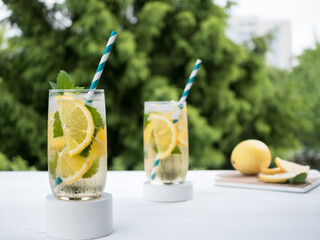 In the garden, on a wooden table, there are glass glasses of lemonade with ice on a plaster podium. Lemon on a cutting board in the background
