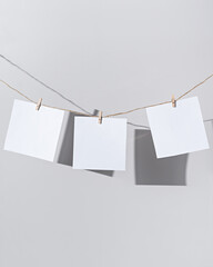Three blank white sheets of square format paper hang from a rope with clothespins. Blank papers on light wall background. Mockup poster with place for your design.