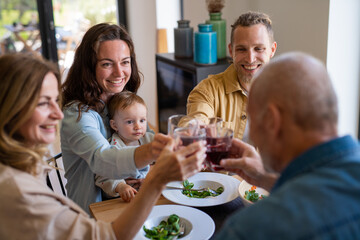 Happy multigeneration family indoors at home eating healthy lunch, clinking glasses.