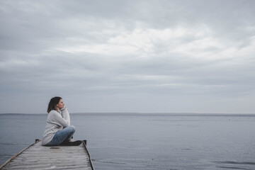 A thoughtful woman in a warm light sweater sits on a wooden pier and enjoys the coolness and fog