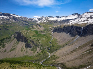 Fototapeta na wymiar Vibrant aerial panoramic of the Alps during springtime. Snow capped peaks, forest covered valley and glacier fed streams and waterfalls