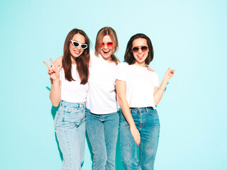 Obraz premium Three young beautiful smiling hipster female in trendy same summer white t-shirt and jeans clothes. Sexy carefree women posing near light blue wall in studio. Cheerful and positive models having fun