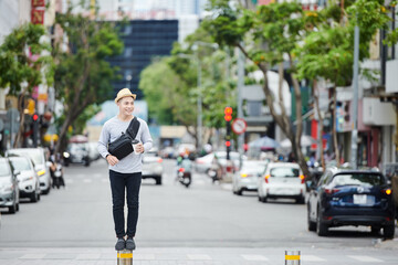 Obraz premium Positive young Asian guy in sun hat standing on small street pole against high traffic and drinking coffee