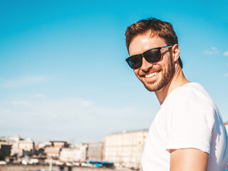 Closeup portrait of handsome smiling  hipster lambersexual model.Stylish man dressed in white T-shirt. Fashion male posing behind blue sky on the street background in sunglasses