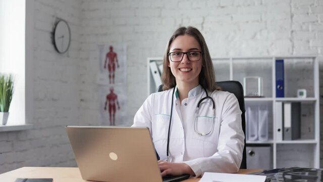 Attractive Caucasian young female doctor in lab coat, glasses and stethoscope over her neck using laptop at desk, looking at camera and smiling while working in medical office. Young medical assistant