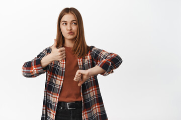 Portrait of indecisive girl shows dislike or like, thumbs up and thumbs down, looking aside...