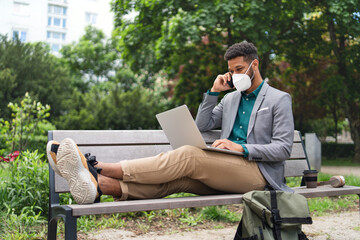 Fototapeta premium Young man with respirator using laptop and smartphone outdoors in city, covid-19 concept.