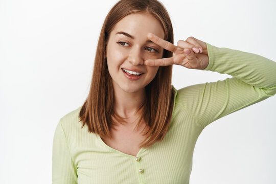 Close up young blond woman in green stylish bloud, show peace v-sign gesture near eye, smiling and looking left at promo beauty text, standing against white background