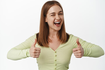 Young cheerful woman winking and showing her support with thumbs up, smiling give confidence boost,...