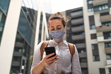 Portrait of young woman with face mask using smartphone outdoors in city, life after covid-19 vaccination.